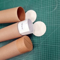 Podłożem zdjęcia jest mata introligatorska, na której znajdują się trzy tuby kartonowe w różnych fazach pakowania wydruku. Od góry tuba pusta z papierowym denkiem, po środku tuba z wystającym plakatem zawiniętym w pergamin i nakleją z logo sklepu, na spodzie tuba zamknięta papierowym denkiem i zabezpieczona taśmą papierową 